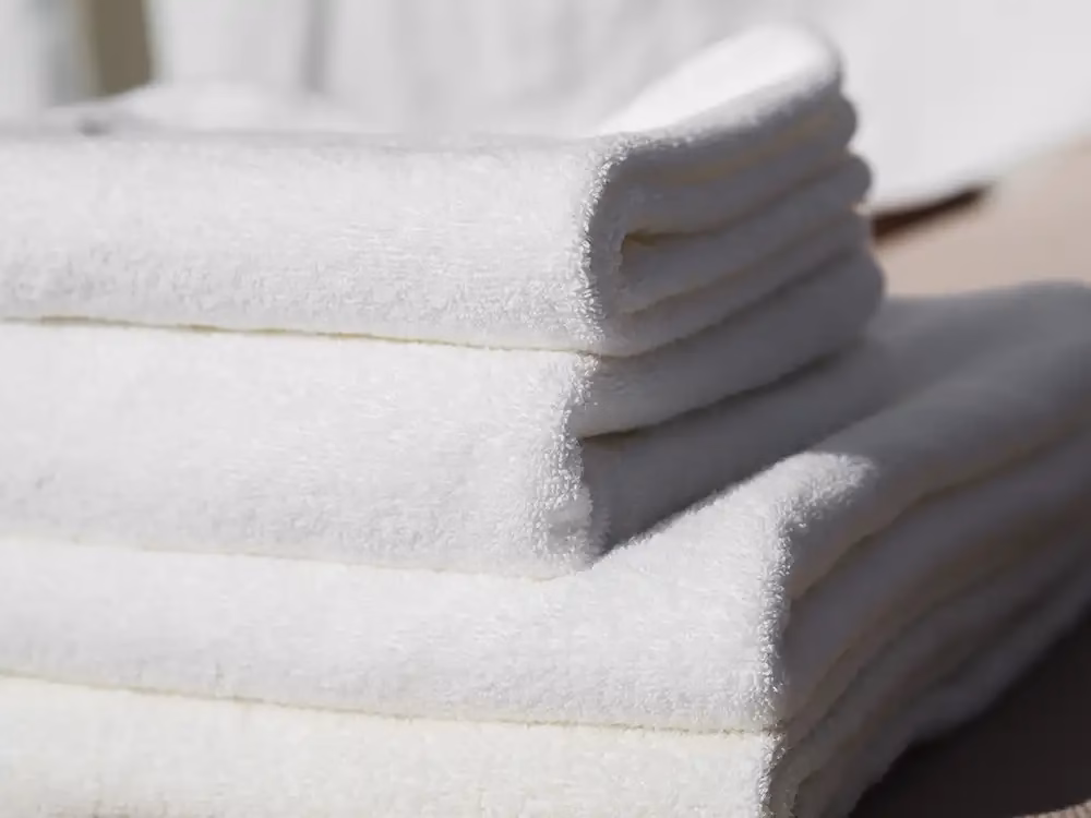 Toalhas brancas técnicas dobradas, limpas e alinhadas em ambiente de hotelaria.
